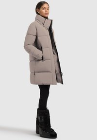 Abrigo tipo puff gris claro con cuello alto, bolsillos con cremallera y un corte ligeramente oversized, combinado con botas negras texturizadas.