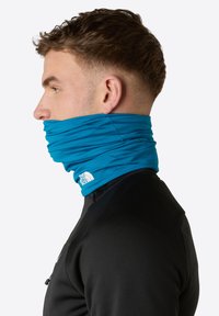 Blå halsduk i elastisk tyg, dragen upp runt halsen. Har en vit logotyp vid basen. Svart topp under.