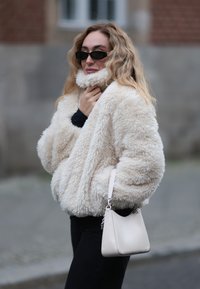 Krém színű, bolyhos műszőrme dzseki magas gallérral, elegáns világos táskával és fekete nadrággal párosítva.