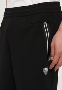 Pantaloni sportivi neri con una finitura testurizzata, dotati di una tasca laterale con zip, due strisce bianche e un piccolo patch del logo EA7.