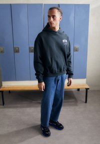 Moletom cinza escuro com bolso canguru e texto branco, jeans azuis, sapatilhas pretas com detalhes azuis, de pé em um vestiário.