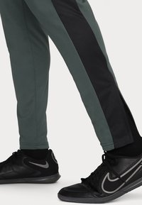 Donkergroene sportbroek met zwarte mesh zijpanelen en ritsdetails, gecombineerd met zwarte sneakers met grijze accenten.