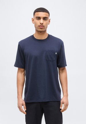 Fiatal férfi áll, tengerészkék, rövid ujjú pólót visel, mellkasi zsebbel, és fekete nadrágot, egyszínű világos háttér előtt.