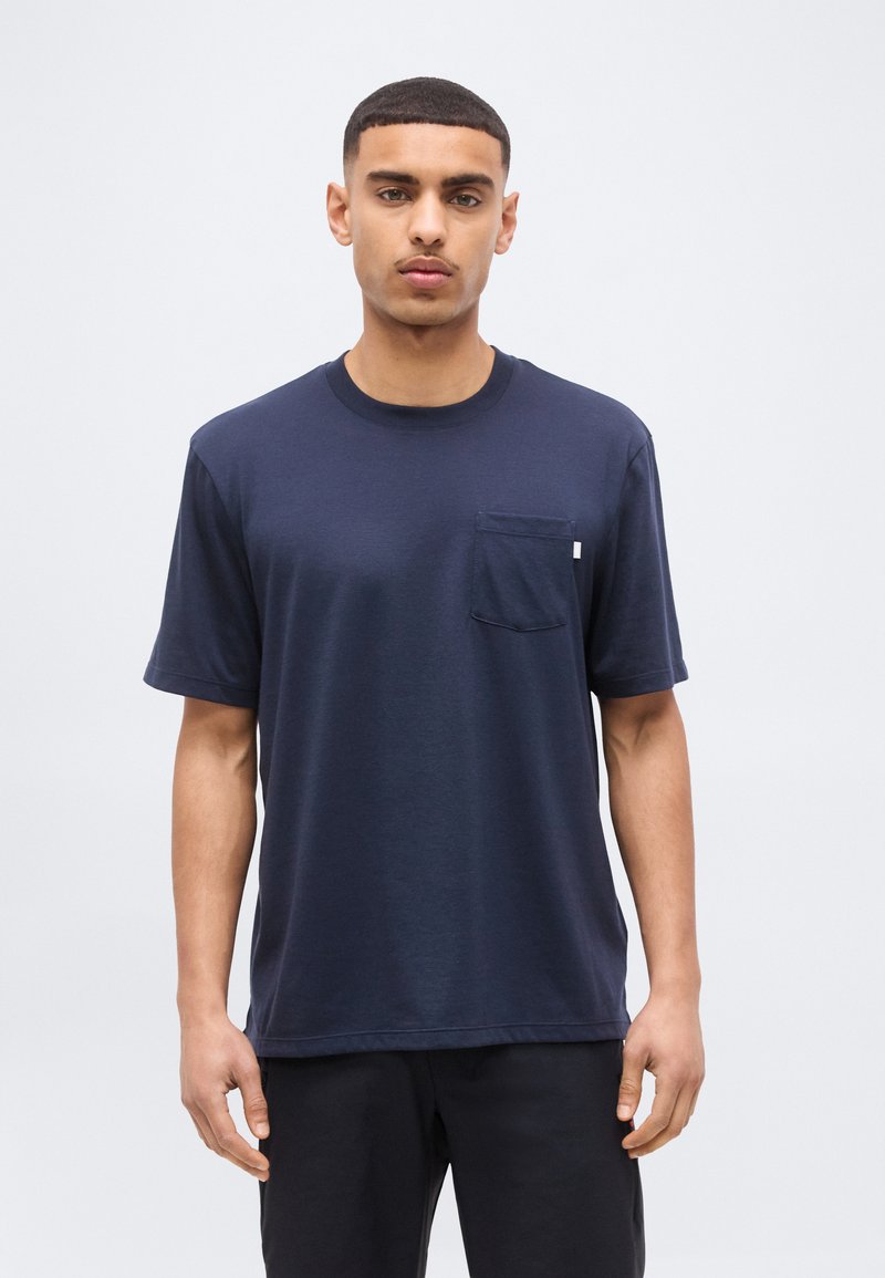 Νέος άνδρας όρθιος, φοράει μπλε ναυτικό κοντομάνικο μπλουζάκι με τσέπη στο στήθος και μαύρο παντελόνι, μπροστά σε απλό φωτεινό φόντο.