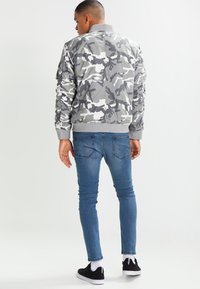 Chaqueta bomber de camuflaje gris con puños y cuello acanalados, combinada con jeans ajustados azules y zapatillas negras.