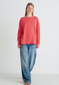 Koraalkleurig longsleeve shirt gecombineerd met lichtblauwe, ruime jeans. Het shirt heeft een ontspannen pasvorm en de jeans hebben een vervaagde textuur.