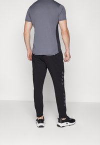 Grå träningströja med kort ärm och svarta sidopaneler, kombinerad med svarta sweatpants med grå chevrondetaljer, bärs med svarta skor.