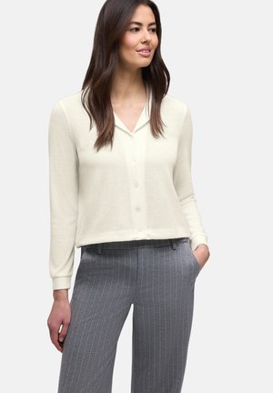 Donna con un cardigan bianco con bottoni e pantaloni grigi a righe sottili, in piedi con una mano in tasca, che guarda leggermente di lato.