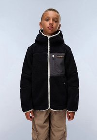 Čierna fleecová bunda s kapucňou, krémovým zipsom a lemom, s šedým náprsním vreckom a textúrovanou látkou. Nosí sa s béžovými nohavicami typu cargo.