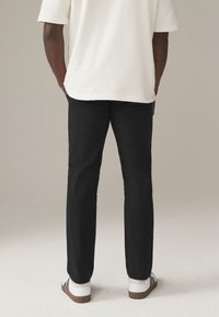 Schwarze Hosen mit geradem Schnitt, kombiniert mit einem weißen T-Shirt mit kurzen Ärmeln und weißen Sneakers mit braunen Sohlen. Minimalistisches Design, keine Muster.
