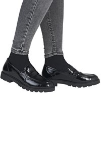 Mocassins en cuir verni noir avec une finition texturée, dotés d'un bout arrondi et d'une épaisse semelle en caoutchouc, associés à un jean gris ajusté.