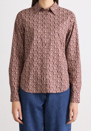 Frau trägt ein langärmliges Hemd mit beige-bordeauxfarbenem Muster und blaue Jeans, steht vor einem schlichten hellen Hintergrund.