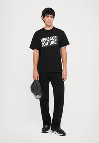 Melna kokvilnas t-kreklu ar "VERSACE COUTURE" logotipu, apvienota ar melniem džinsiem, tumšu cepuri un melniem sporta apaviem ar baltiem akcentiem.