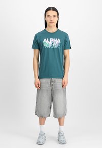 Tyrkysové bavlněné tričko s grafikou "ALPHA INDUSTRIES" v barevných vlnitých písmenech. Vybaveno šedými denimovými šortkami a světle šedými vzorovanými teniskami.