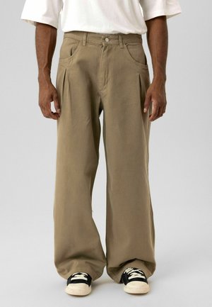 Kaki wijd uitlopende broek van zachte stof met voorplooien, zijzakken en een knoopsluiting, gecombineerd met zwart-witte sneakers.