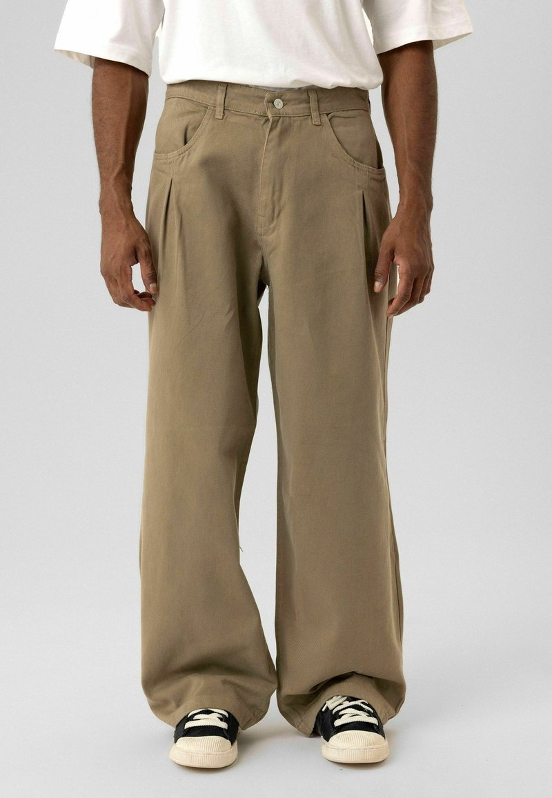 Kaki wijd uitlopende broek van zachte stof met voorplooien, zijzakken en een knoopsluiting, gecombineerd met zwart-witte sneakers.