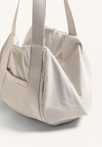 Ljust beige tyg-shoppingväska med en mjuk, strukturerad design. Har breda remmar och en sidficka som framhäver dess funktionella stil.