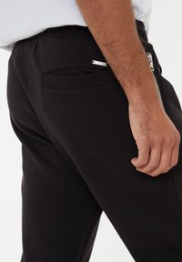 Schwarze Jogginghosen aus strukturiertem Stoff, mit einer seitlichen Reißverschlusstasche und einer normalen Gesäßtasche an der rechten Hüfte.