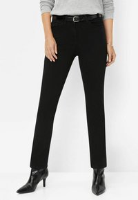 Pantalons en denim noirs à jambes droites, dotés d'une ceinture avec une boucle circulaire, de poches avant et portés avec des talons aiguilles noirs.