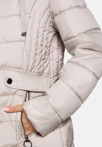 Doudoune beige clair avec texture matelassée, poche zippée, rabat à boutons-pression et poignets élastiqués aux manches.