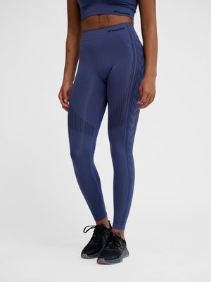 Fit person iført blå højtaljede leggings med teksturerede side mønstre og sorte atletiske sko, stående mod en ensfarvet baggrund.