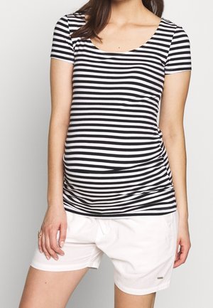 Nainen, joka käyttää mustavalkoista vaakaraitakuosista lyhythihaista paitaa ja valkoisia shortseja, seisoo yhdellä kädellä reidellä.