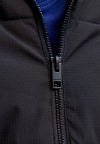 Melna dūraiņu jaka ar izteiktu rāvējslēdzēju, kas aprīkots ar zīmolu nesošu piesaistes cilpiņu, pārklāj zilu kreklu. Virsma ir gluda ar smalku tekstūru.