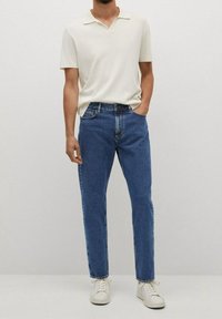 Hombre vestido con una camiseta polo blanca de manga corta, jeans azules y zapatillas blancas, de pie con una mano en el bolsillo frente a un fondo liso.