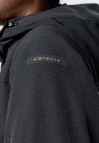 Zbliżenie na rękaw czarnej kurtki z przeszytym prostokątnym naszywką z logo marki "ICEPEAK".