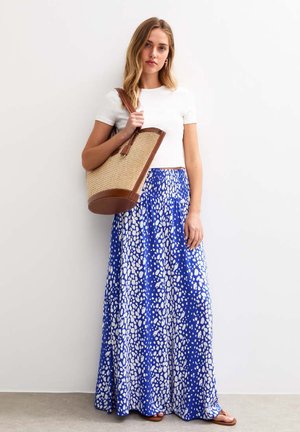 Maxi-jurk in blauw met witte stippen, gecombineerd met een eenvoudige witte top. Geweven rieten handtas met bruine leren accenten. Bruine sandalen.