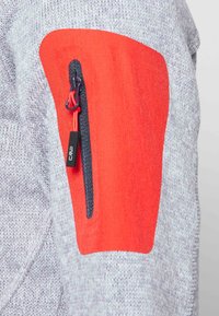 Grijze gebreide kleding met een helder oranje accent op de elleboog en een met rits afgesloten zak met een zwarte trekband. Textuurdetails van de stof zijn zichtbaar.
