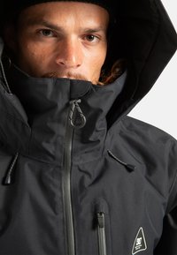 Czarna wodoodporna kurtka z wysokim kołnierzem, regulowanym kapturem i zamykanymi na zamek kieszeniami. Gładka faktura z refleksyjnymi akcentami.