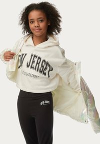 Gekürzter cremefarbener Hoodie mit "NEW JERSEY" Aufdruck, kombiniert mit schwarzen Leggings. Das Model trägt eine schillernde Jacke.