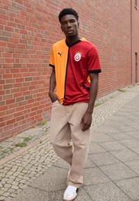 Galatasaray fodboldtrøje med orange og kastanje sektioner, sort kant, med logo og tre stjerner. Sammen med beige bukser og hvide sneakers.