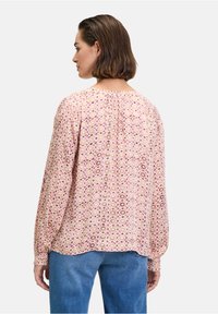 Blusa a maniche lunghe in tessuto rosa chiaro con motivi circolari in crema e viola. Presenta uno scollo posteriore arricciato e polsini elasticizzati.