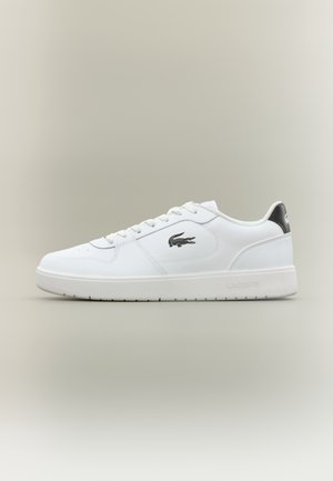 Hvid lavtop sneaker med snøredesign, sort krokodillelogo på siden og sort hælstrop, vist på beige baggrund.