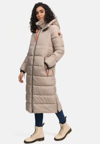 Beige long puffer coat with a hood, featuring horizontal quilted design, contrasting orange accents, and front zippered pockets. Paired with beige boots.