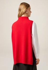 Rotes ärmelloses Weste mit hohem Kragen, geripptem Saum und weicher Textur, kombiniert mit weißem Langarmshirt und schwarzem Rock.