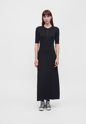 Une femme porte une robe noire longue jusqu’aux chevilles, à manches courtes et boutonnée sur le devant, avec des baskets montantes noires, sur un fond uni.