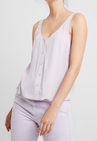 Light lavender sleeveless top with a V-neck, front button closure, and slightly flared hem. Smooth texture and minimalist design.