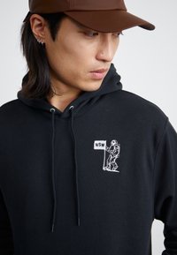 Hoodie noir avec cordon de serrage, avec un graphique d'astronaute brodé blanc et "NSW" sur la poitrine. Tissu doux, coupe décontractée.