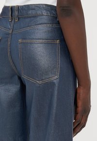 Donkerblauwe, hoge taille denim jeans met een glanzende textuur, gele stiksels en twee achterzakken met een kenmerkende vorm.
