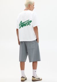 Witte oversized t-shirt met groen graffiti-stijl logo op de achterkant, gecombineerd met grijze shorts, witte sokken en beige/zwarten sneakers.
