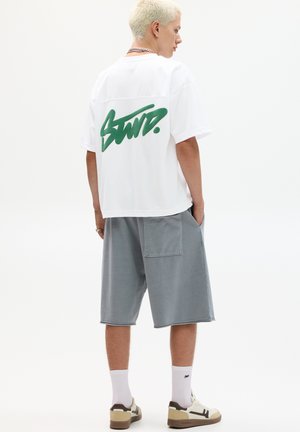 White oversized t-shirt with green graffiti-style logo on the back, paired with gray shorts, white socks, and beige/black sneakers.