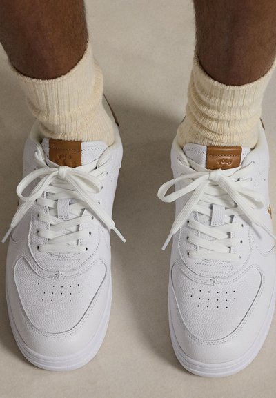 Picioare purtând șosete crem cu rebru și adidași albi din piele, cu pete maro din piele pe limbă, văzute de sus.