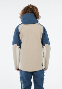 Heren buitenjas in beige en marineblauw, met een blauwe capuchon, ritssluitingen aan de zijkanten en een ontspannen pasvorm. Gemaakt van weerbestendig materiaal.