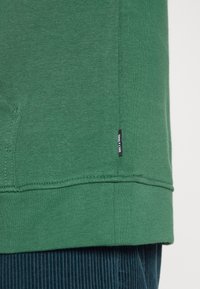 Sudadera de algodón verde con un dobladillo acanalado, presenta una pequeña etiqueta de marca negra en el lateral. Tela texturizada, corte casual, sin patrones visibles.