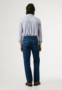 Jeans de mezclilla azul con corte recto, combinados con una camisa de rayas clara, vistos por la parte de atrás, con bolsillos y un parche de cuero en la cinturilla.