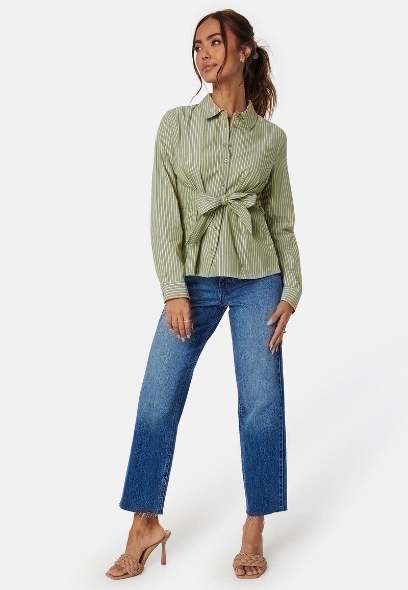 Grün-weiß gestreifte Bluse mit geknotetem Vorderteil. Kombiniert mit hochgeschnittenen blauen Jeans und cremefarbenen Riemchen-Heels.