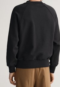 Person in einem schwarzen Langarm-Sweatshirt mit gerippten Bündchen und Saum, kombiniert mit braunen Hosen, von hinten gezeigt.